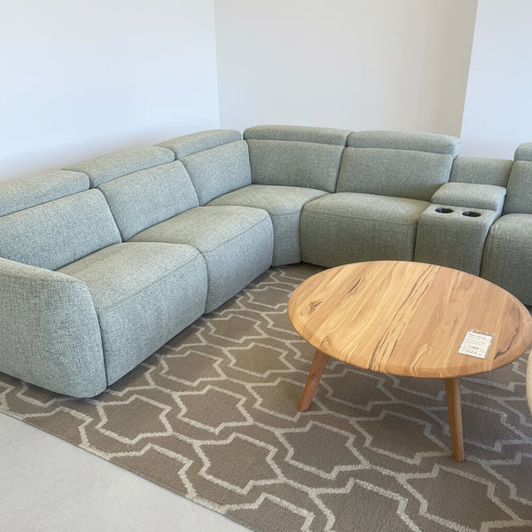 A light sage green modular sectional sofa in a furniture showroom, paired with a round natural wood coffee table and a gray geometric patterned rug.