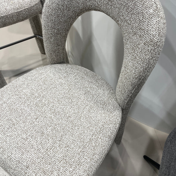 A close-up of a modern upholstered chair with a curved, looped backrest and a rounded seat cushion, both covered in a textured light gray woven fabric.