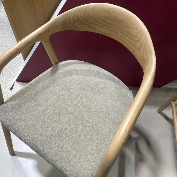 A close-up of a modern wooden chair with a curved light oak frame and a light gray upholstered seat cushion, shown against a dark red and white background.