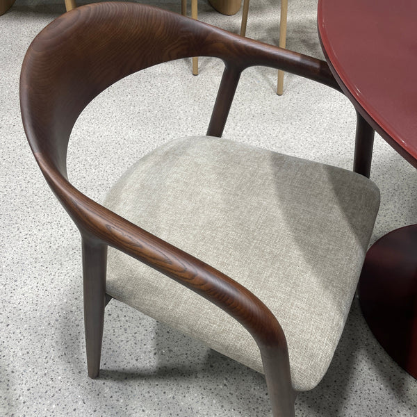A modern dining chair with a curved dark brown wooden frame and a light beige textured fabric seat cushion, shown on a speckled gray floor.