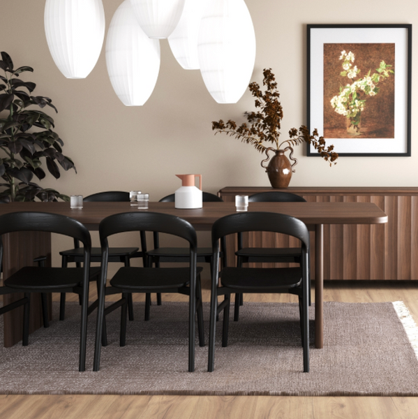 A styled dining room interior featuring a dark wood dining table with black chairs, a matching wood sideboard, white pendant lights, a gray rug, and a framed floral art print on the beige wall.