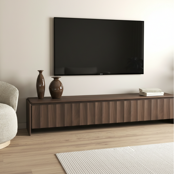 A tall framed floral art print with white lilies on a brown background, displayed leaning against a beige wall above a dark wood sideboard. The room includes a gray upholstered chair on the left, two brown vases, and a small stack of books on the sideboard, with a plant on the right.