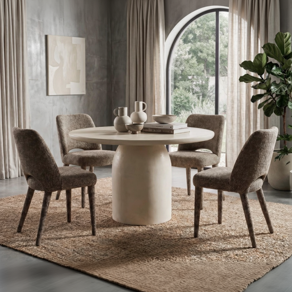 A round dining table with a thick pedestal base and four upholstered dining chairs in a warm neutral-toned dining room. The set is styled on a textured jute rug with a concrete wall, arched window, and potted plant in the background.