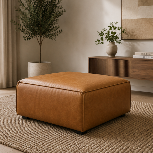 A square tan leather ottoman with a smooth, slightly glossy finish, styled in a modern living room setting on a textured beige rug next to a wooden sideboard and potted plants.