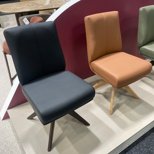 Three modern upholstered accent chairs in black, tan, and sage green, arranged on a white display riser in a showroom.