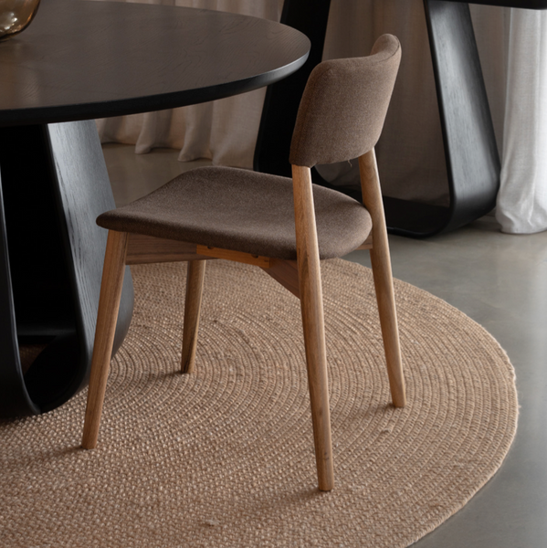 A modern dining room scene featuring a light brown upholstered wooden chair in the foreground, set on a round woven jute rug next to a dark round table. In the background, a black console table with a white decorative vase stands against a backdrop of beige curtains.
