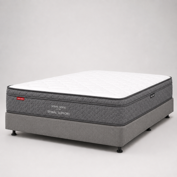 A mattress on a gray upholstered support base, photographed in a clean studio setting with a white background. The mattress has a white quilted top and dark gray sides with visible text reading 'SPINAL SUPPORT' and a small red logo tag.