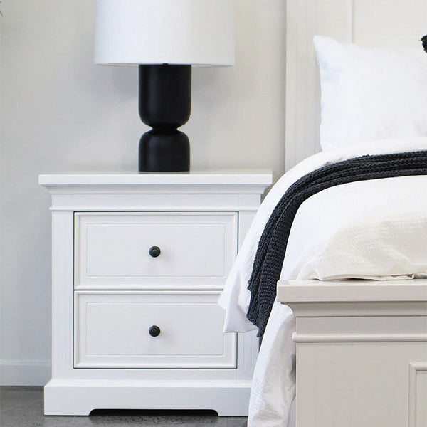 White bedside nightstand with drawers and black knobs beside the bed