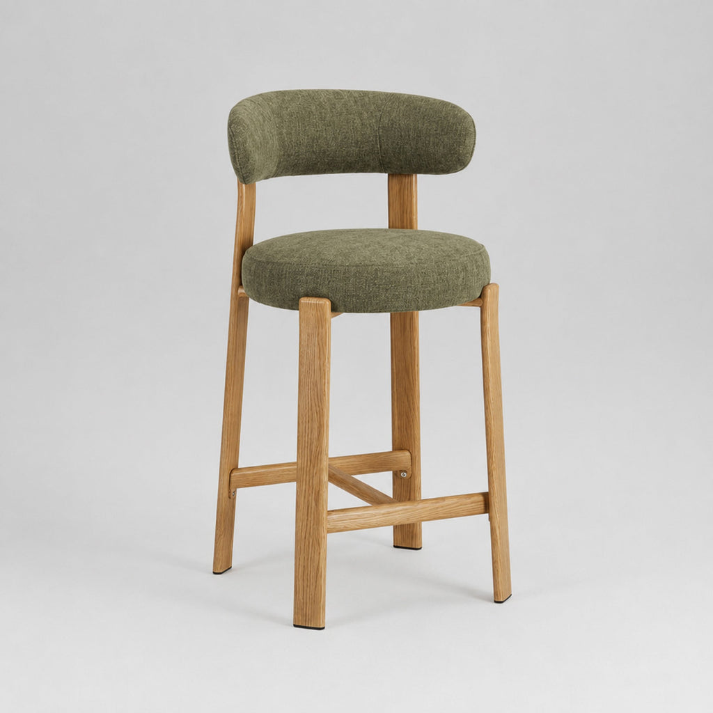 A modern bar stool with a curved olive green upholstered backrest and seat cushion, supported by a light natural wood frame with cross-bracing, shown on a light gray studio background.