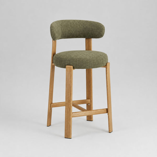 A modern bar stool with a curved olive green upholstered backrest and seat cushion, supported by a light natural wood frame with cross-bracing, shown on a light gray studio background.