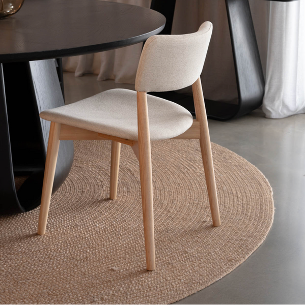 A modern dining chair with a light beige upholstered seat and backrest, supported by light natural wood legs, positioned on a round woven jute rug next to a dark round table.